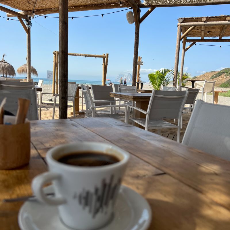 Tavolo in legno con tazzina da caffè, dehors sul mare e vista lontana sulla Rocca di Gibilterra.